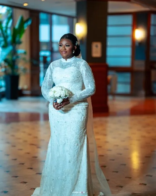 Beautiful bride in long-sleeved white lace wedding gown holds white bouquet while posing in elegant hotel lobby.