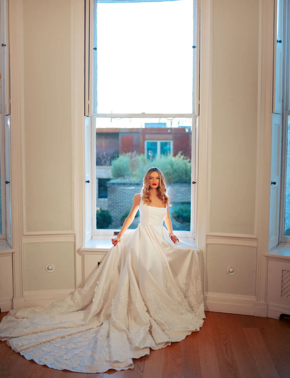 Elegant strapless wedding gown with long train photographed in front of a tall window in a bright room.