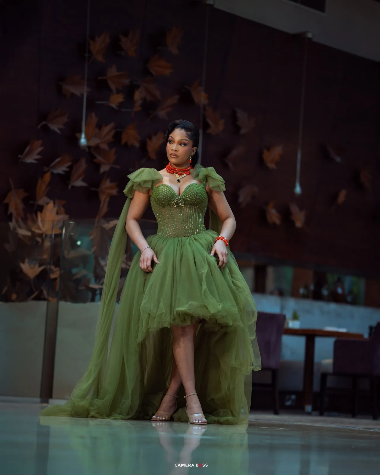 A person poses in an elegant olive green high-low evening gown with puff sleeves and tulle skirt on a stage.