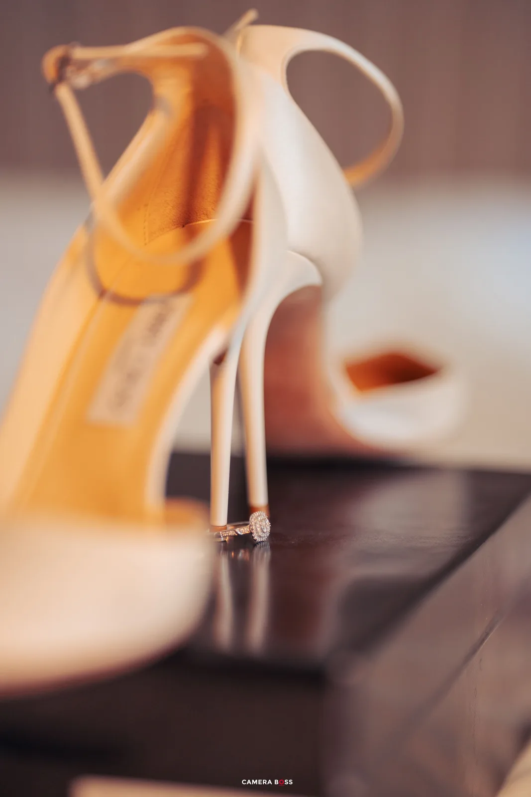 An elegant white stiletto heel with ankle strap sits atop a dark surface, with an amber-colored shoe blurred in background.
