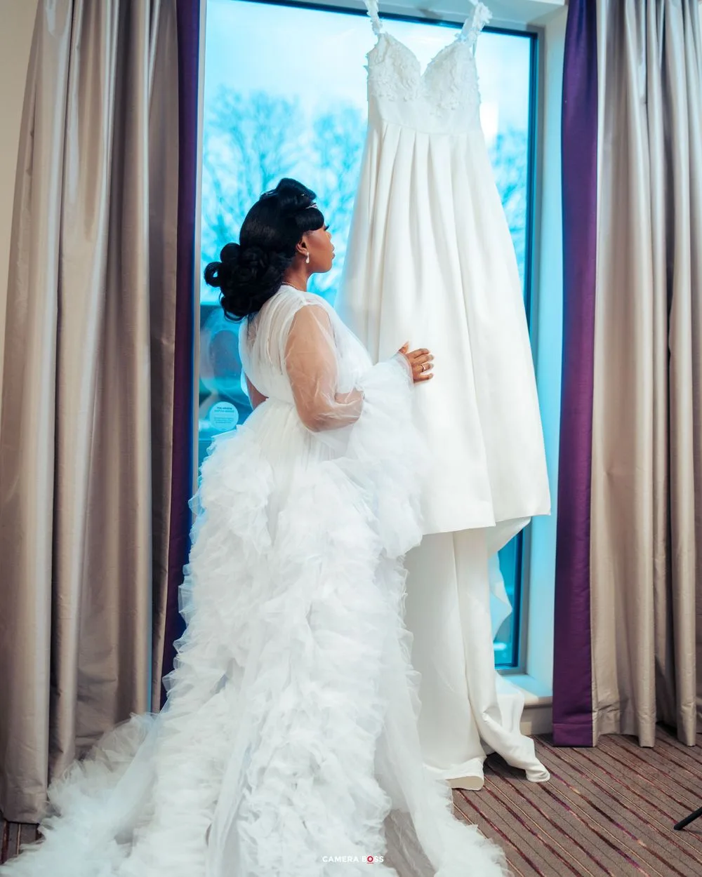 A figure in a white ruffled gown stands looking at a classic white wedding dress displayed by a window.