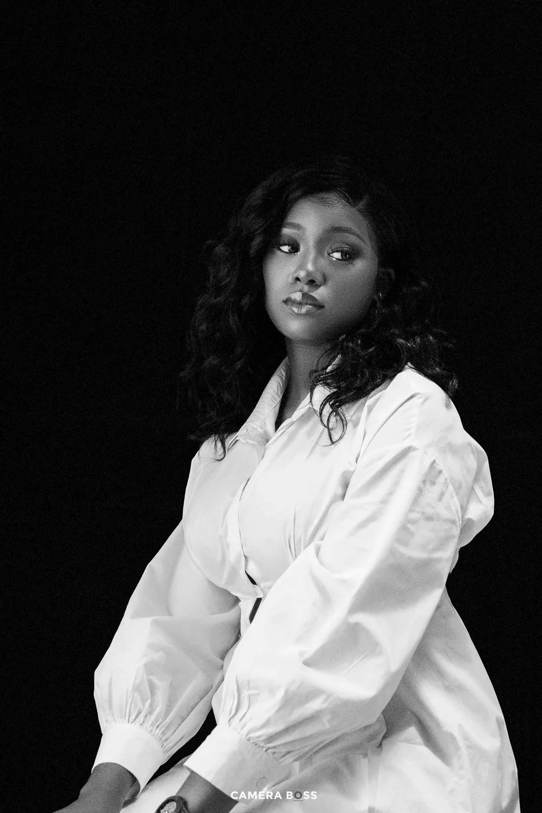 A black and white portrait of someone wearing a white button-down blouse with puff sleeves against a dark background.