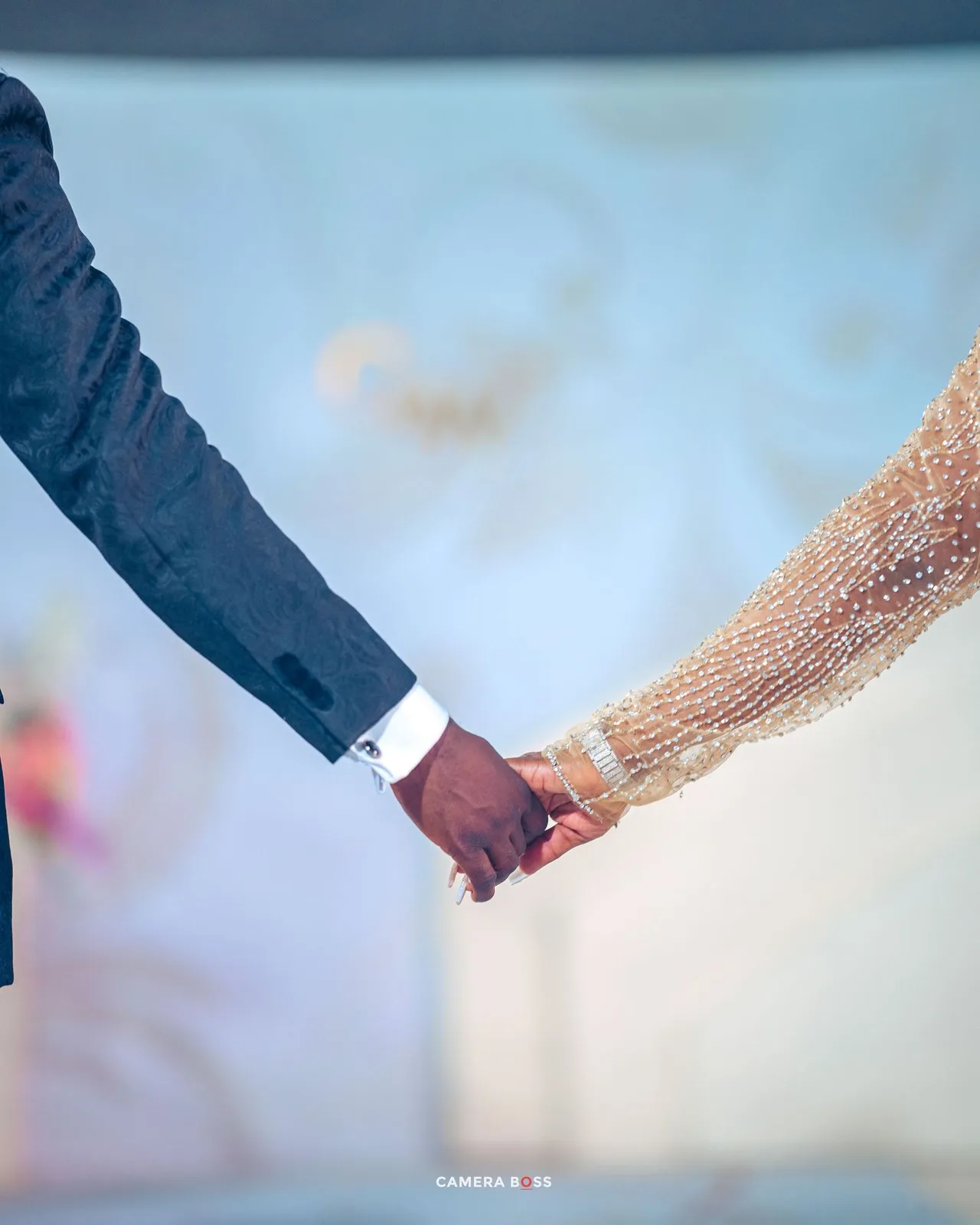 A close-up shot of two people holding hands, one wearing a dark suit sleeve and the other in sparkly formal attire.