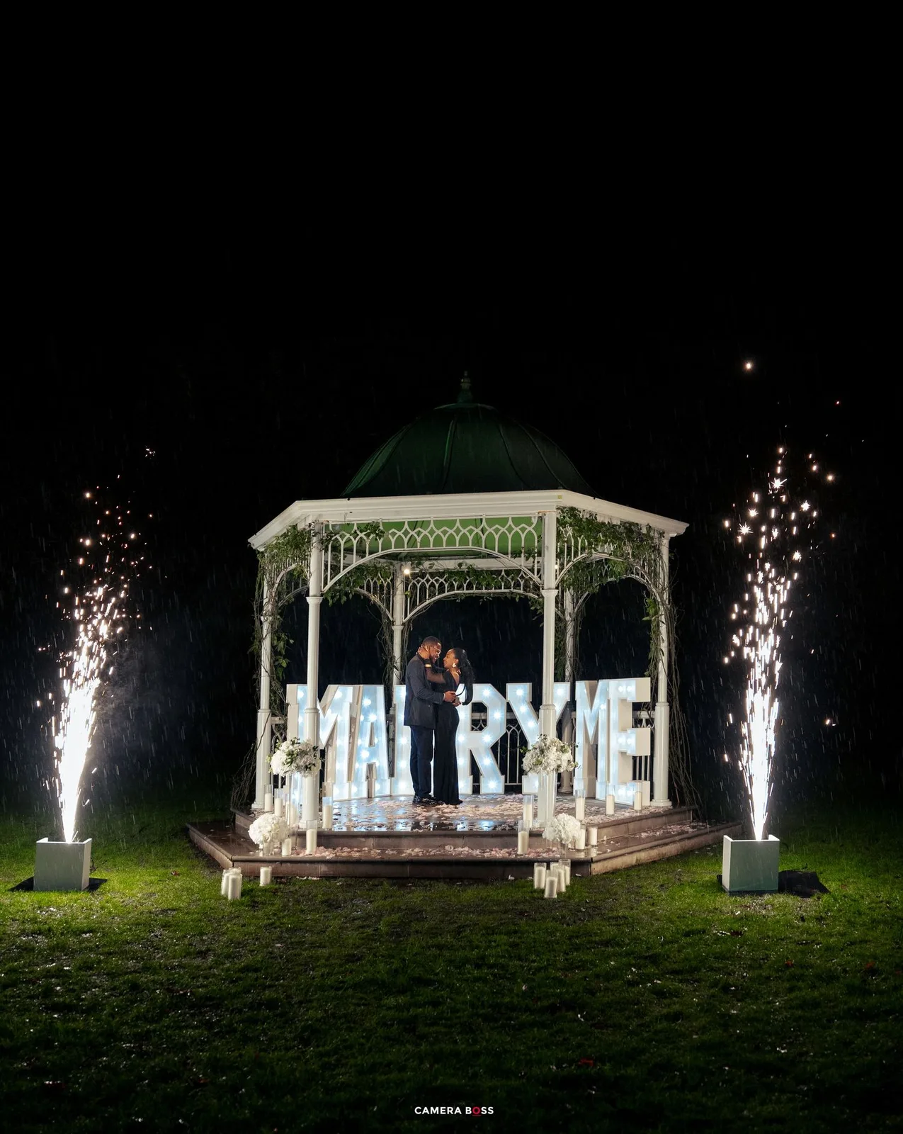 A white wedding gazebo illuminated at night with large sparklers on both sides and lit-up MARRY ME letters on display.