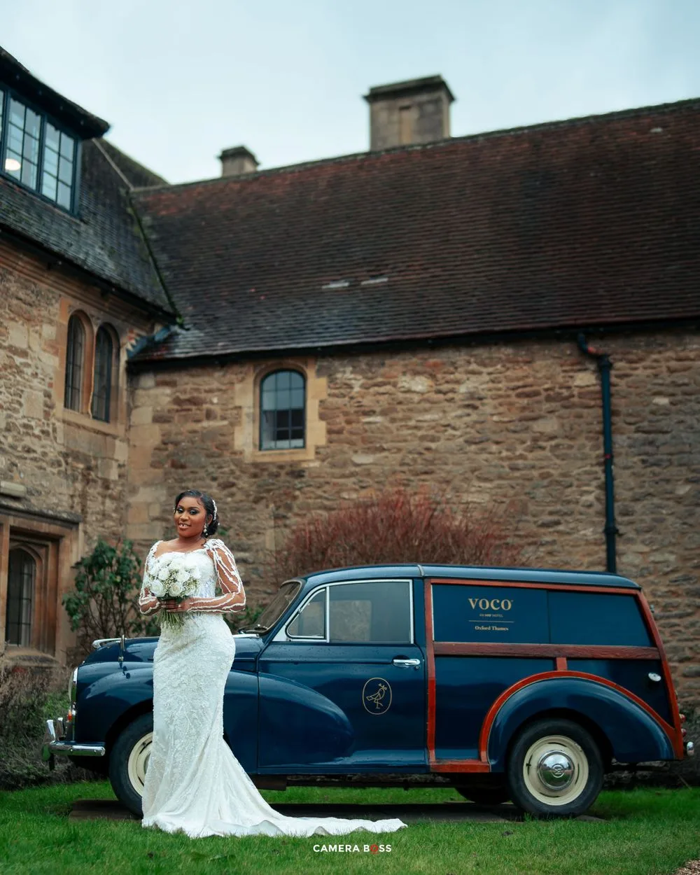 A white lace wedding gown stands elegantly next to a vintage blue delivery van against a stone building backdrop.