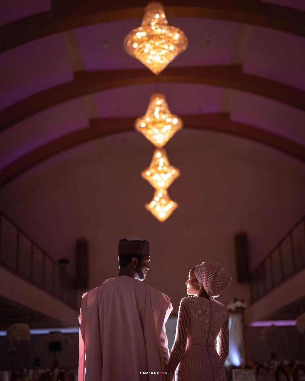 A couple in traditional dress stand beneath illuminated chandeliers creating a romantic silhouette in an elegant venue.