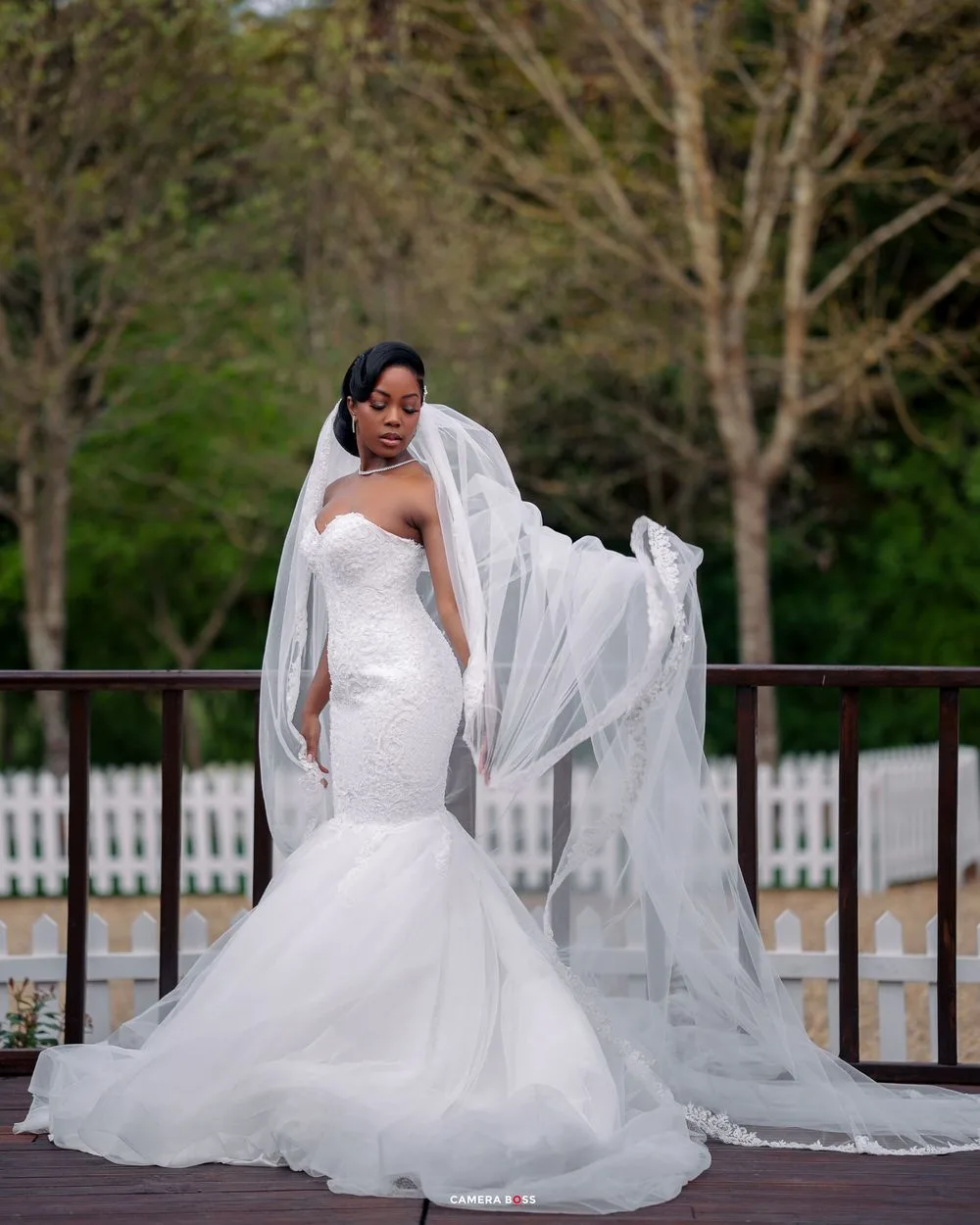 A stunning bridal portrait in a white wedding gown against a rustic wooden fence with trees in the background.
