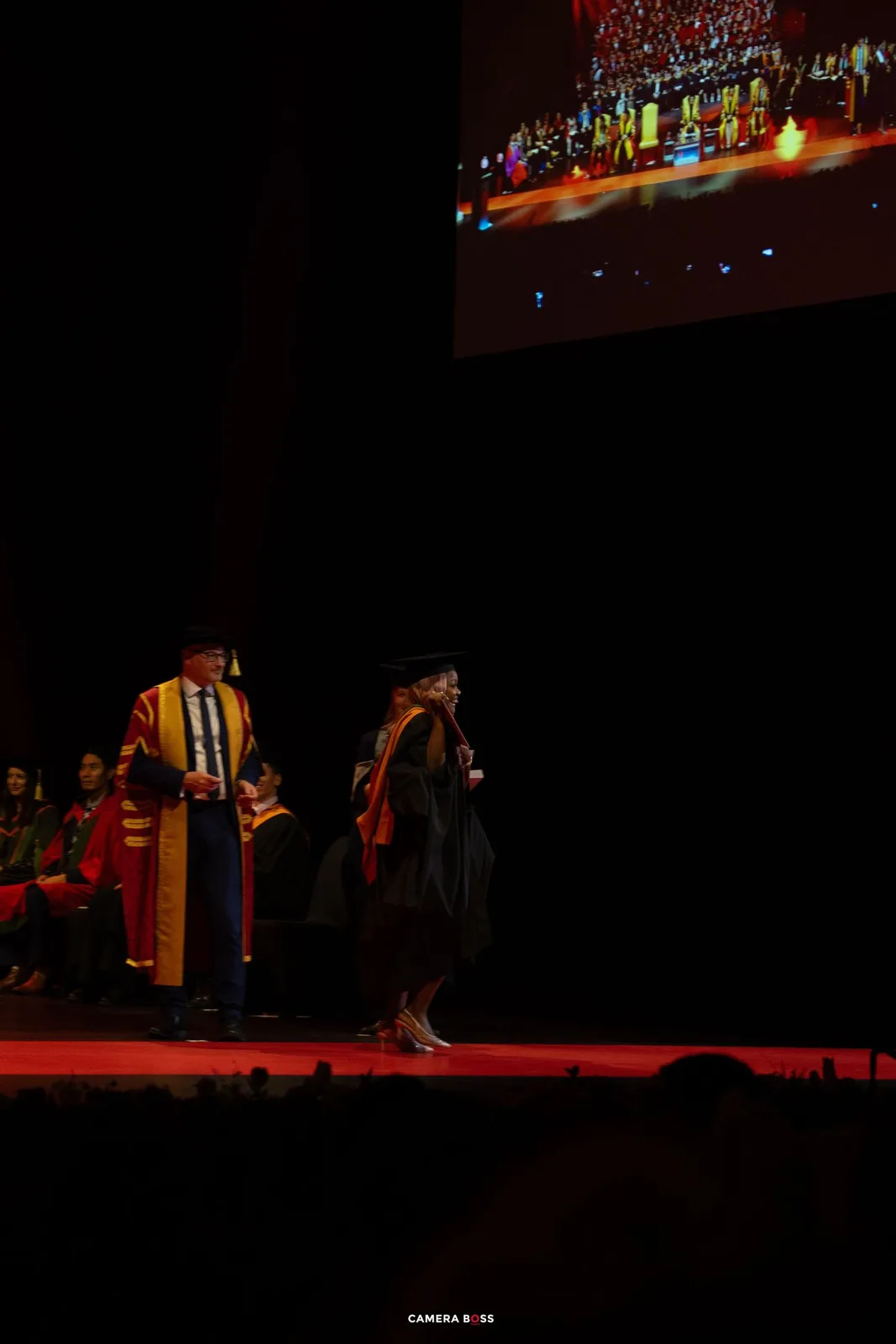 Graduates in academic regalia walk across a dimly lit stage during a commencement ceremony with red lighting effects.
