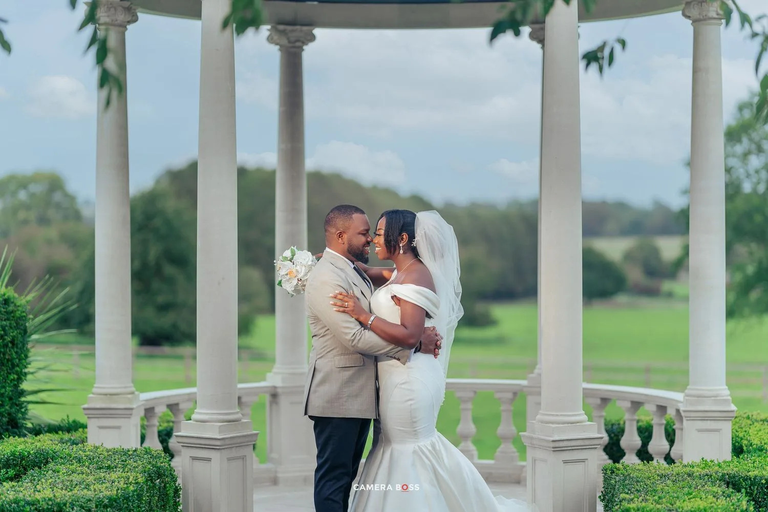 A couple shares a romantic moment under an elegant white gazebo surrounded by lush green landscaping.