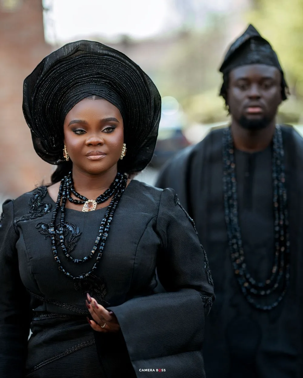 Traditional Nigerian couple wearing black attire with gele headwrap and beaded necklaces at formal ceremony.