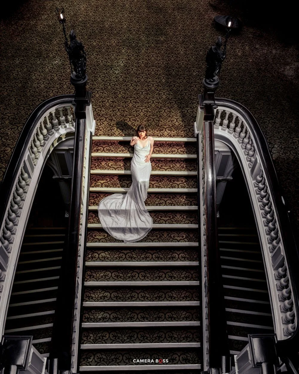 A bride in a flowing white dress stands gracefully on an ornate grand staircase with elegant curved railings and dark lighting.