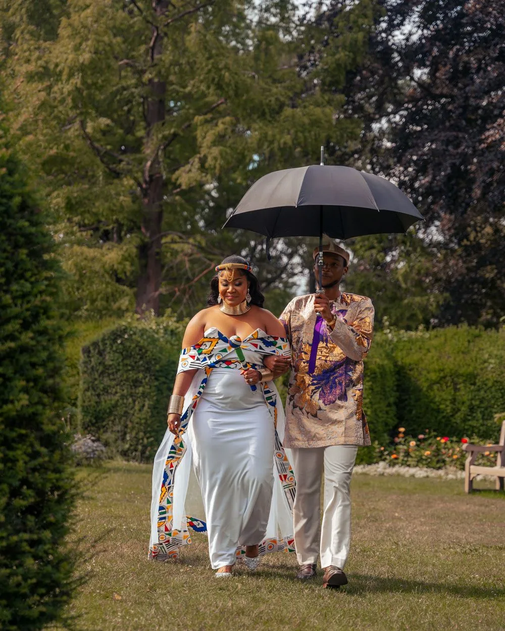 Two people in colorful floral attire walk together under a black umbrella in a garden setting during a wedding ceremony.