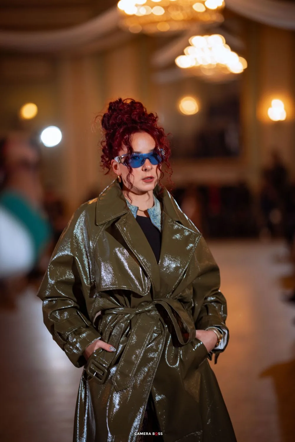 A person wearing a dark olive patent leather trench coat and blue sunglasses walks with dramatic burgundy hair styled up.