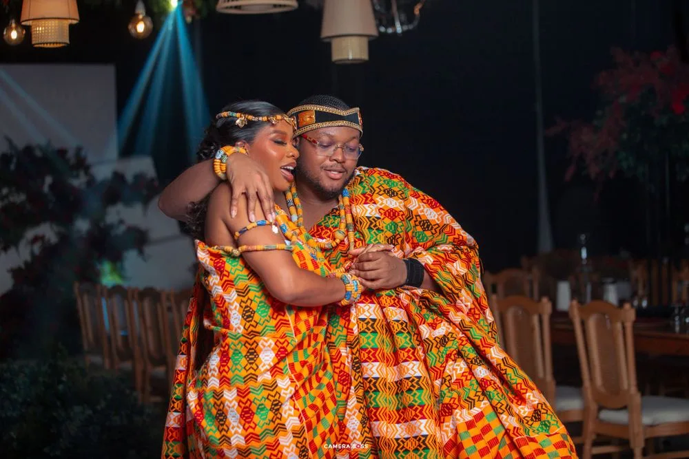 Two people wearing vibrant orange and yellow traditional African kente cloth embrace while smiling during a celebration.