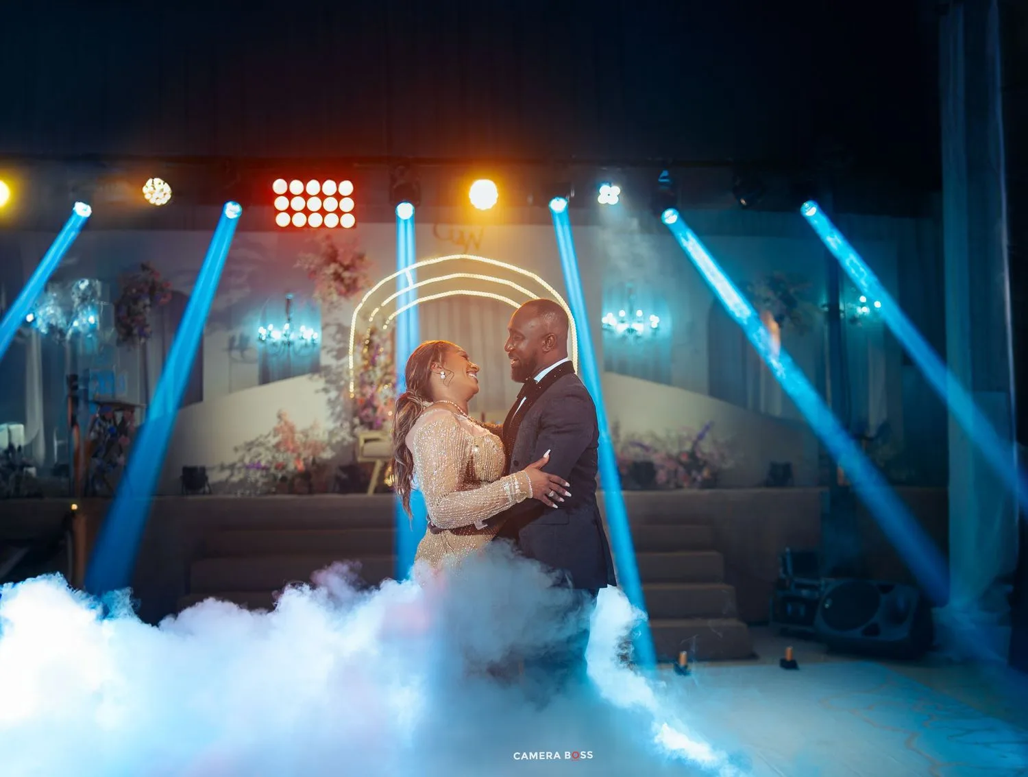 A couple shares their first dance on a misty dance floor illuminated by dramatic blue and orange stage lighting.