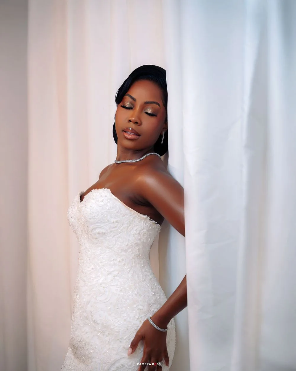 A bride poses elegantly against a white curtain backdrop in a fitted white lace strapless wedding gown.