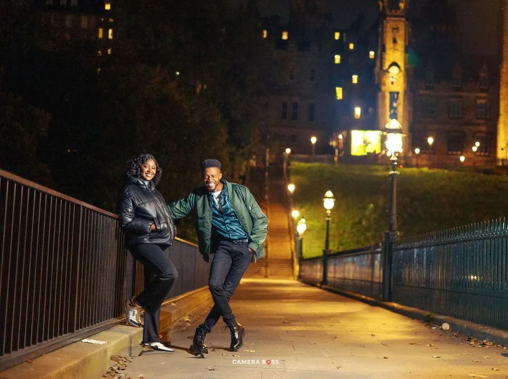 Two people lean against railing on illuminated path at night with city lights and tower in background.