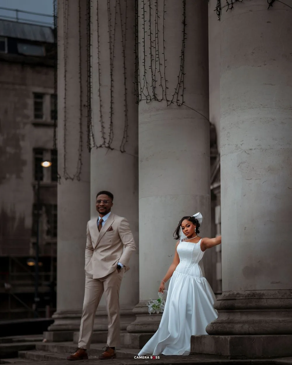 A couple poses elegantly between white classical columns at dusk, with the bride in a flowing white dress and groom in beige suit.