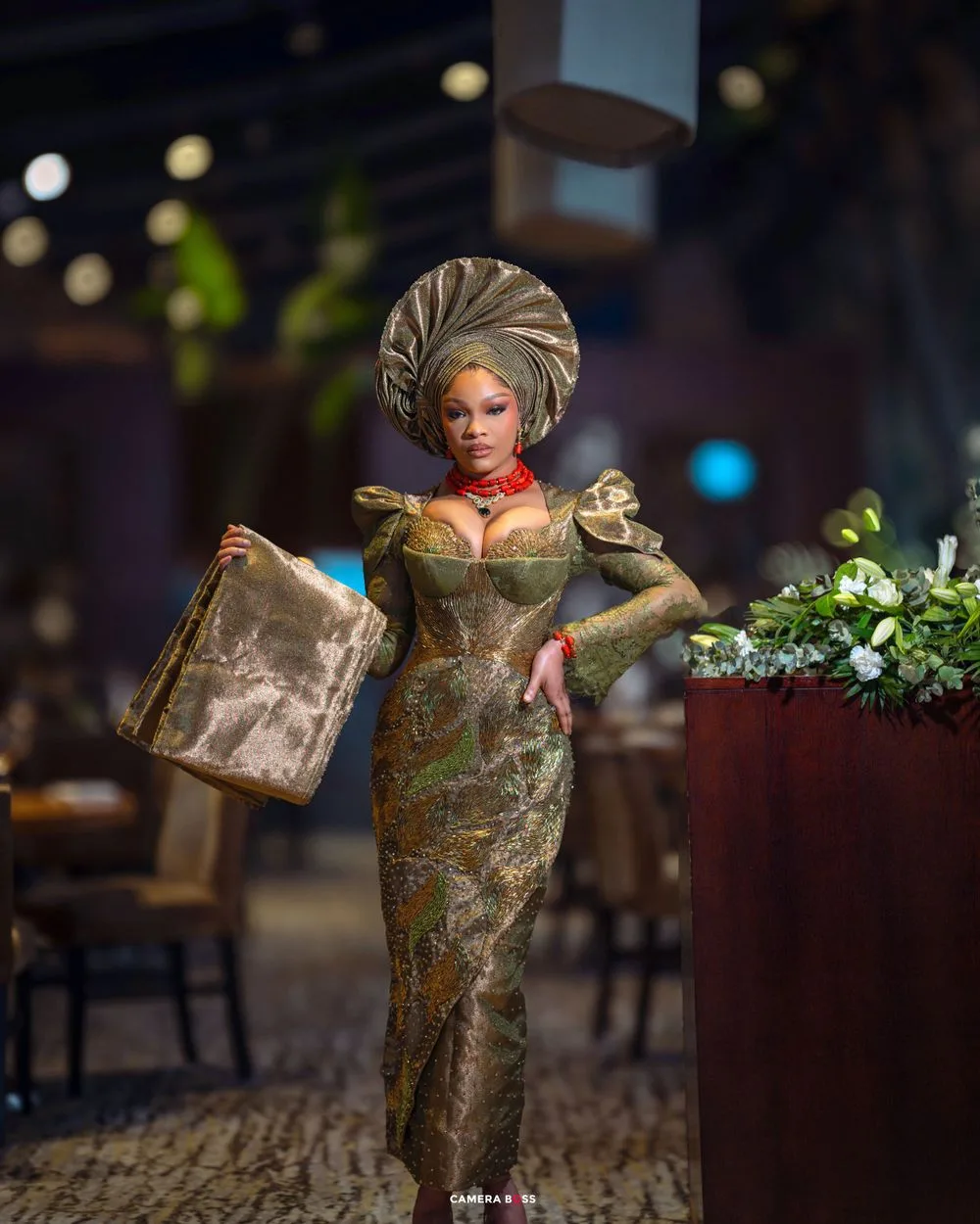 A woman in an elegant green traditional dress and turban headpiece poses with a decorative bag in an upscale restaurant setting.