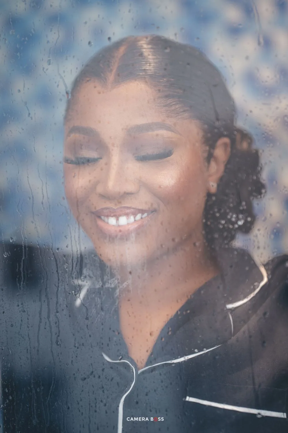 A dreamy portrait through rain-speckled glass shows someone wearing a black jacket with white piping, eyes closed and smiling serenely.