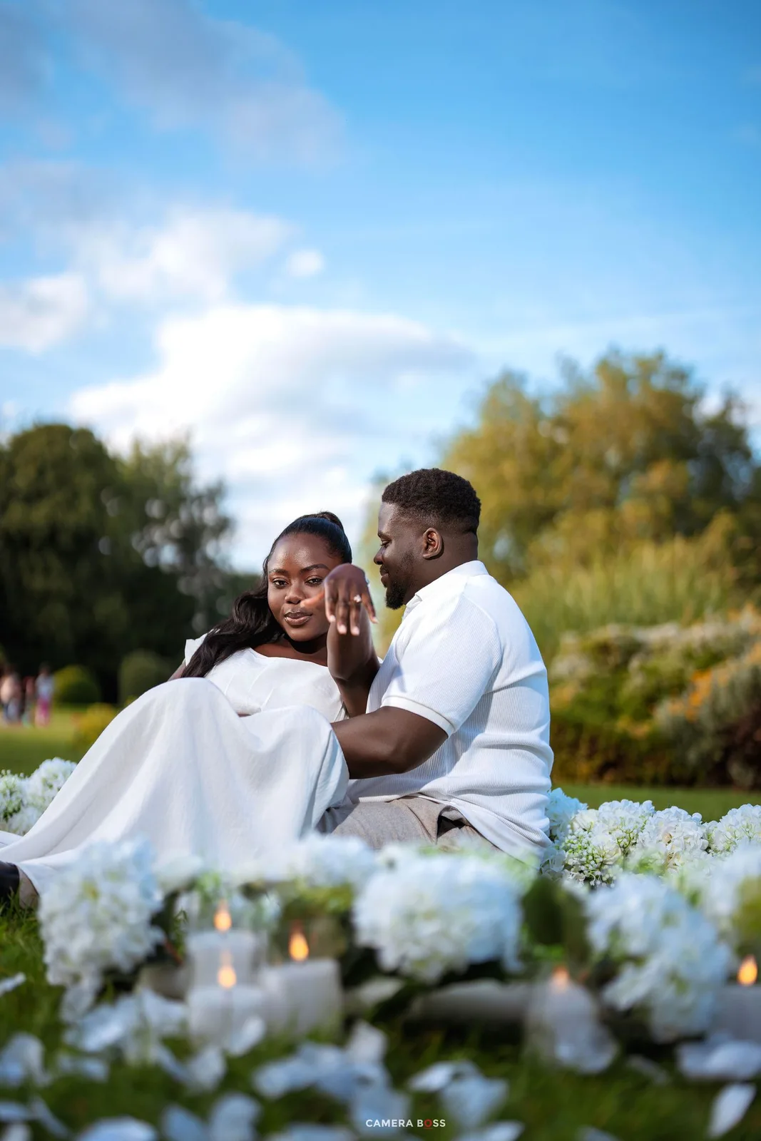 Couple in white outfits sharing a romantic outdoor proposal moment surrounded by white flowers and candles on green grass.