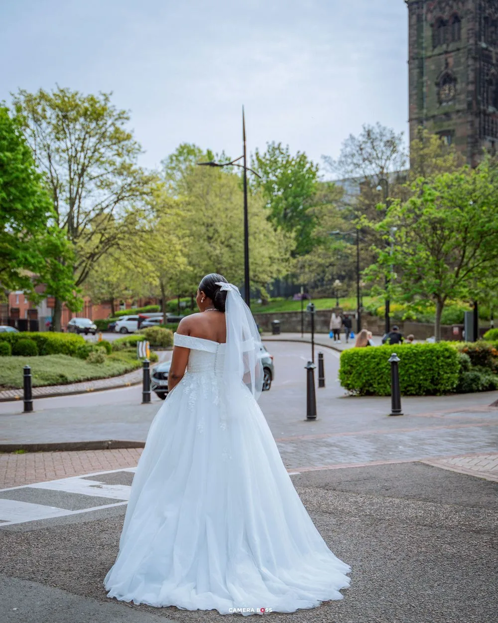A bride in a white off-shoulder wedding gown and veil stands in front of a historic church with stone architecture.