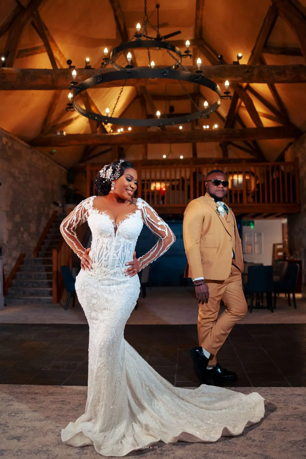 A stylish couple poses in a rustic venue with exposed beams and hanging lights, wearing formal wedding attire.