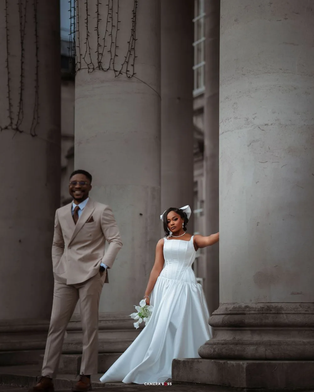 A couple in wedding attire poses joyfully between elegant white columns of a classic building.