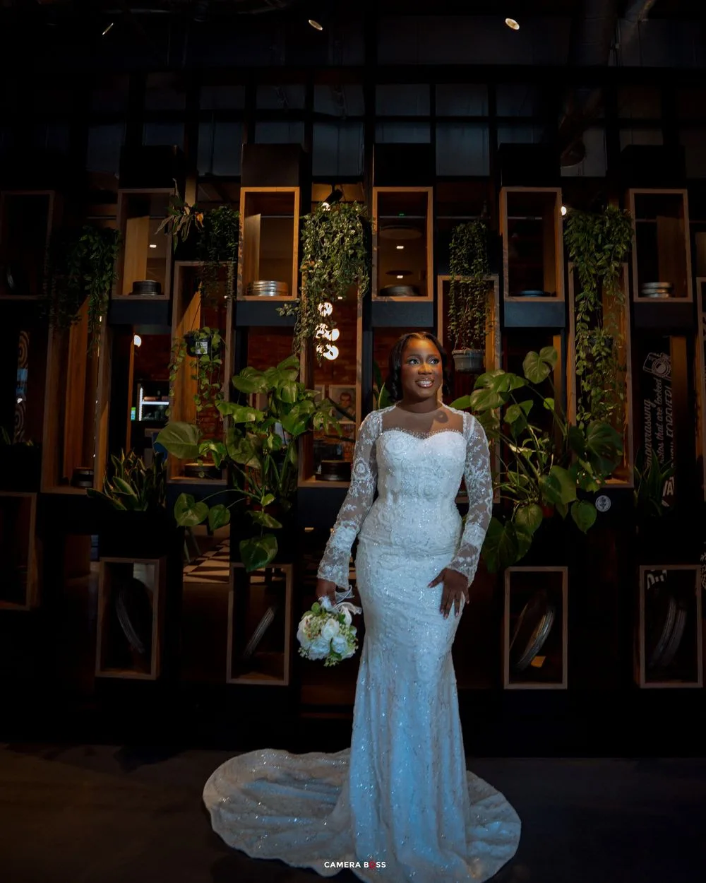 A bride in a white lace gown poses elegantly in front of a dark background with illuminated windows.