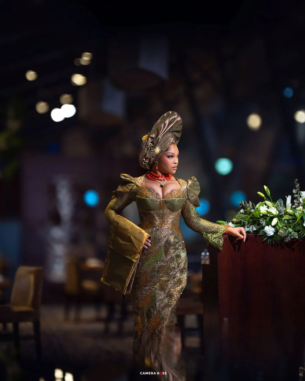 A glamorous figure in a gold brocade dress and elaborate feather hat poses dramatically in a dimly lit setting with bokeh lights.