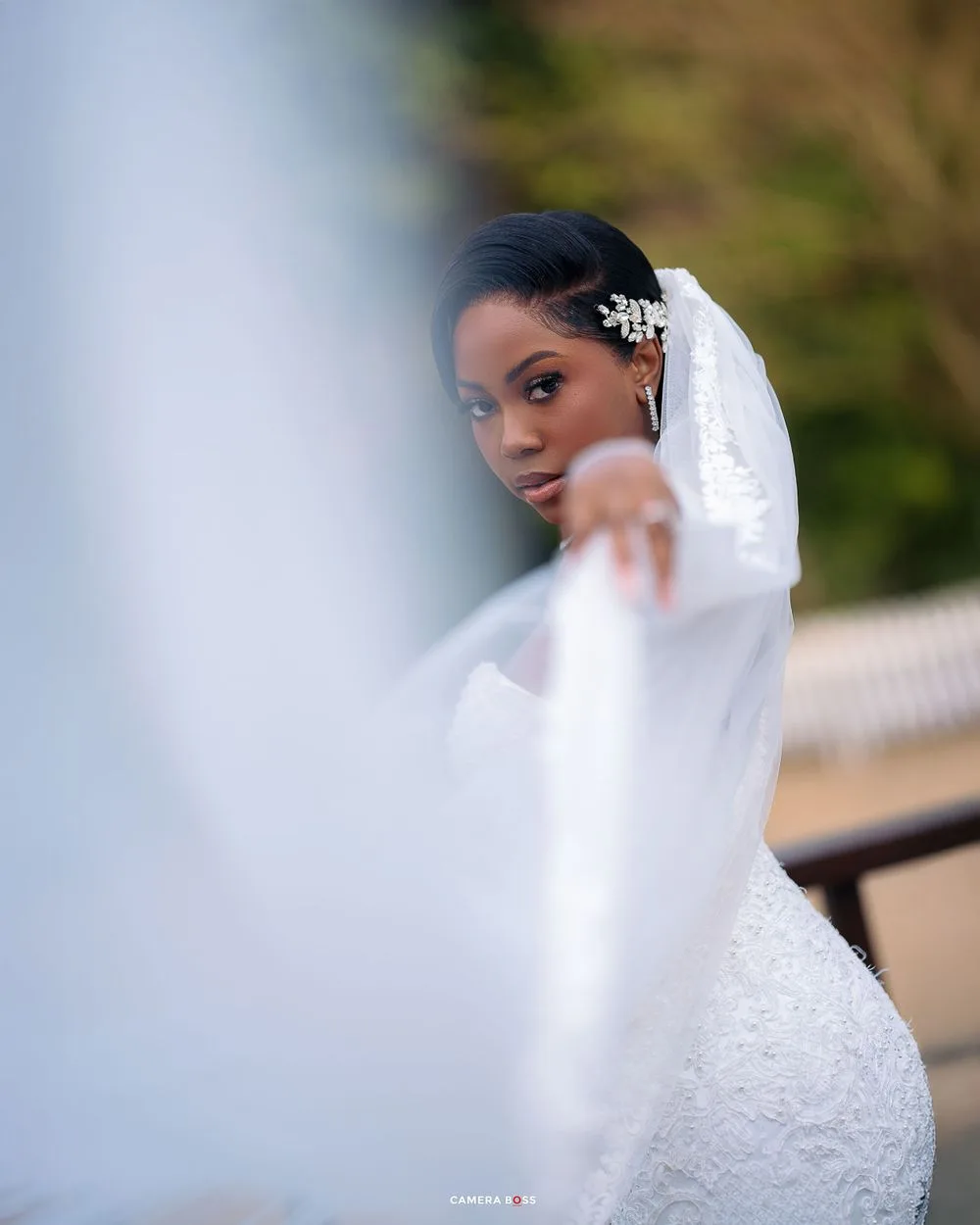 A bride in an elegant white lace wedding dress and veil poses against a soft blurred background.