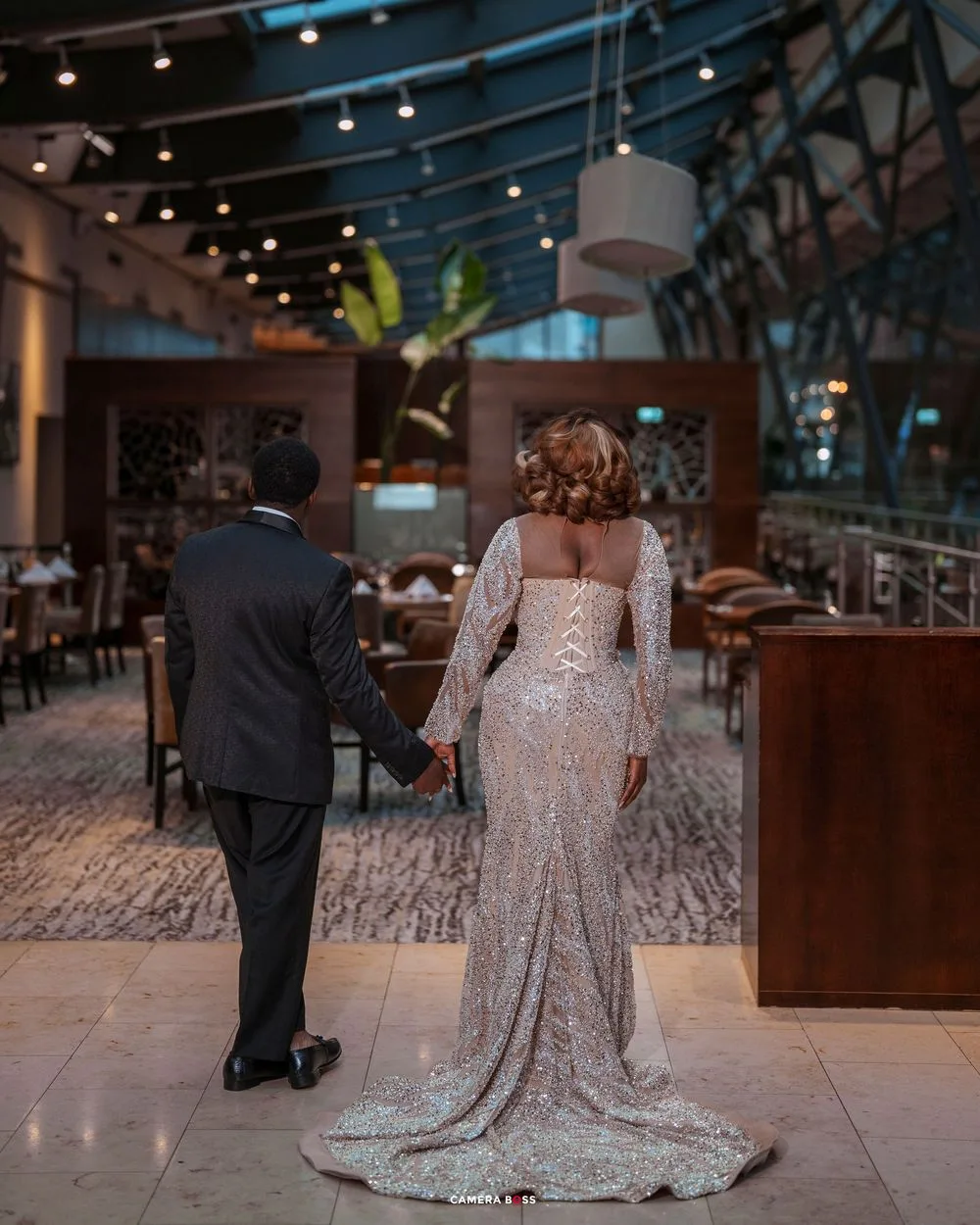 Couple holding hands in elegant venue, woman in sequined gown with train, man in dark suit, romantic wedding photo.