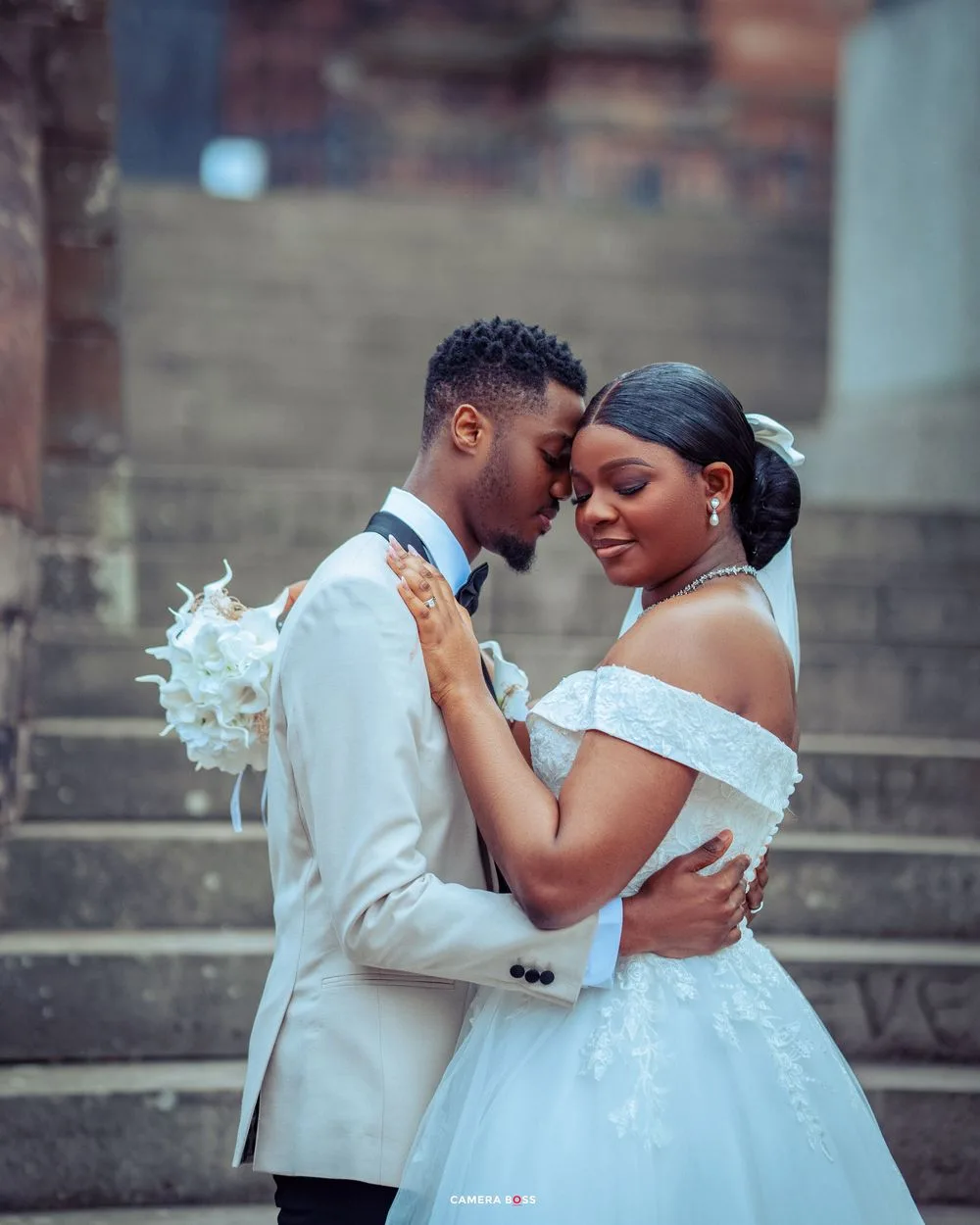 A romantic wedding photo featuring a couple in white formal attire embracing on stone steps.