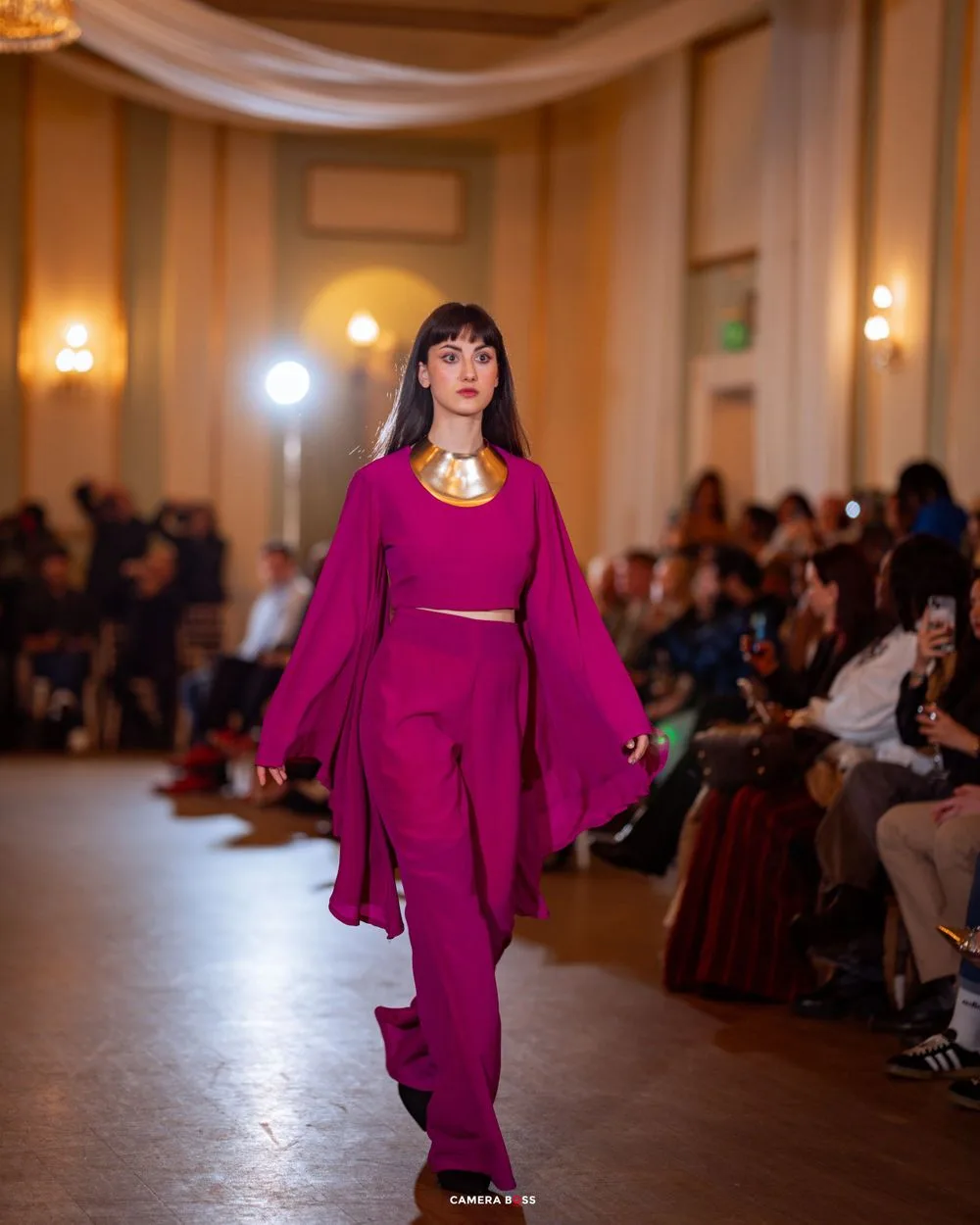 A model walks the runway in a vibrant fuchsia ensemble with dramatic flowing sleeves during a fashion show.