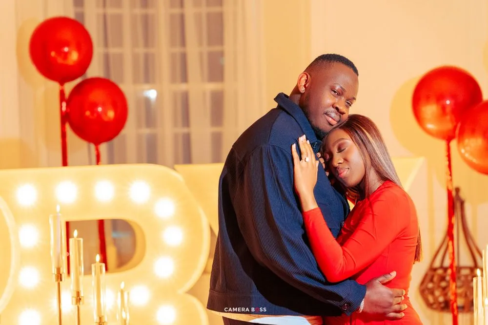 A romantic couple embraces in front of illuminated letter lights and red balloons during a special moment.
