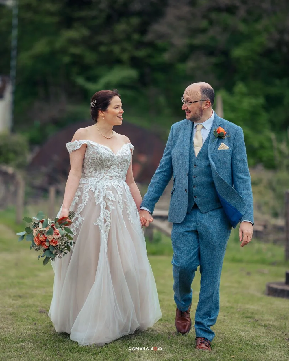 A couple on their wedding day walks hand in hand outdoors, with the bride in white and groom in blue.