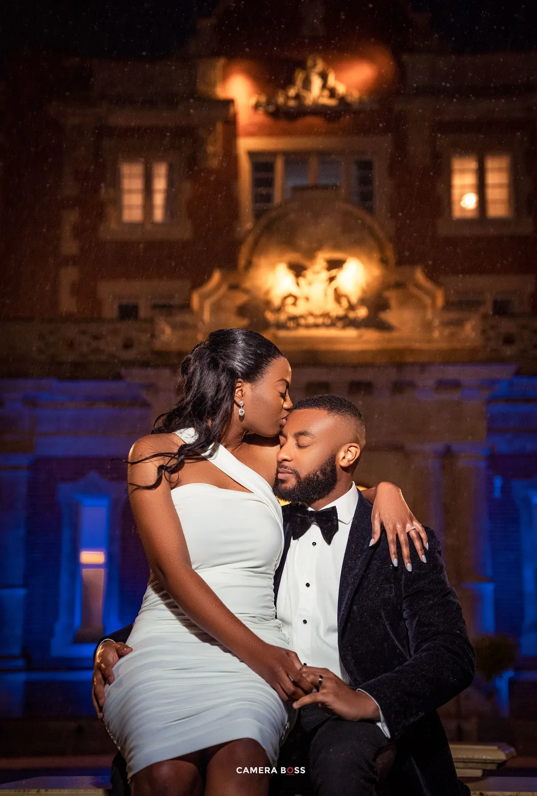 A romantic evening portrait of a couple in formal wear against an illuminated historic building.