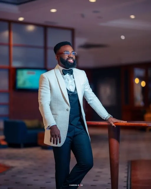Stylish figure in white tuxedo jacket and black bowtie poses confidently in dimly lit venue with ambient lighting.