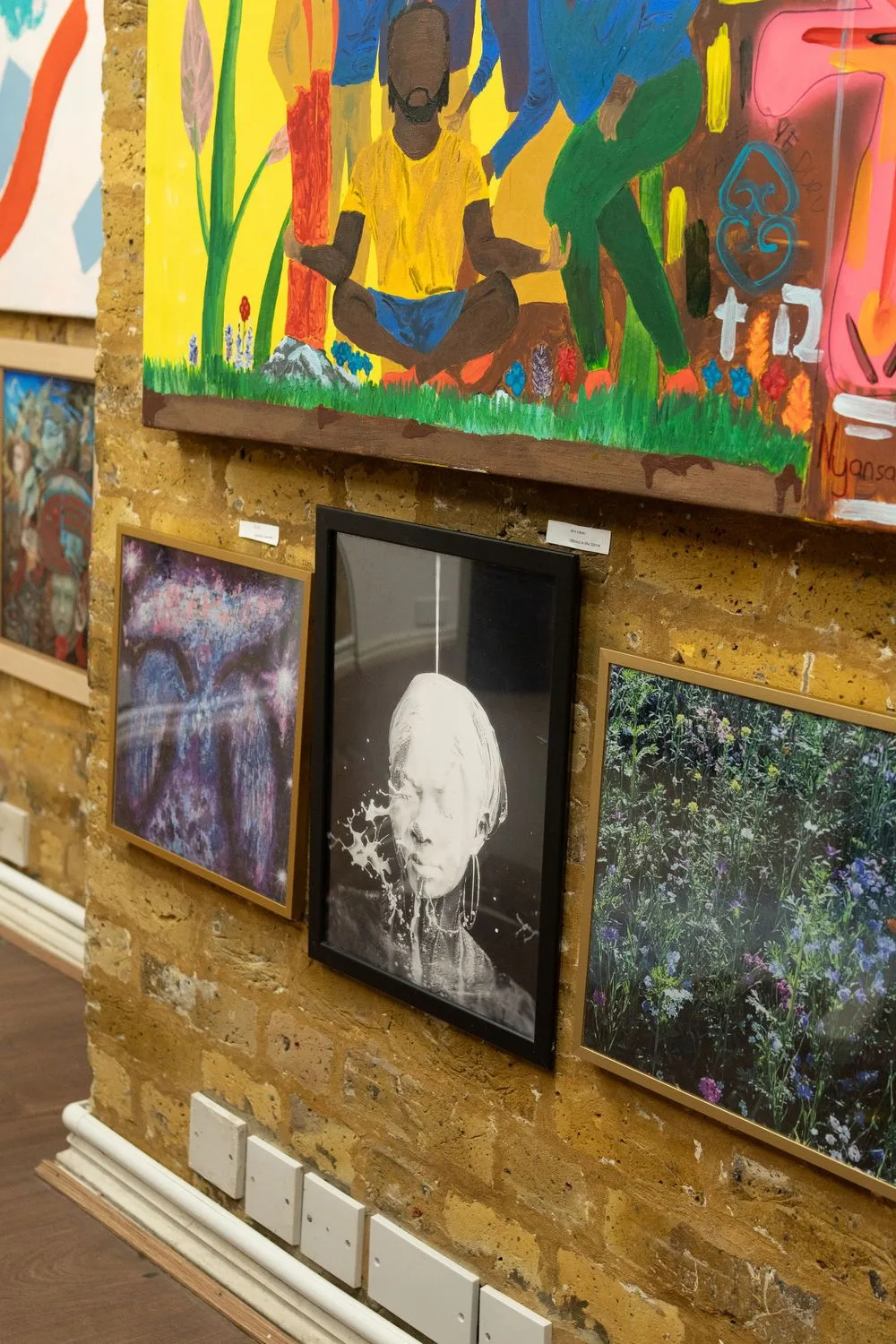 Art gallery wall displaying colorful mural paintings and black and white photography on rustic wooden backdrop.