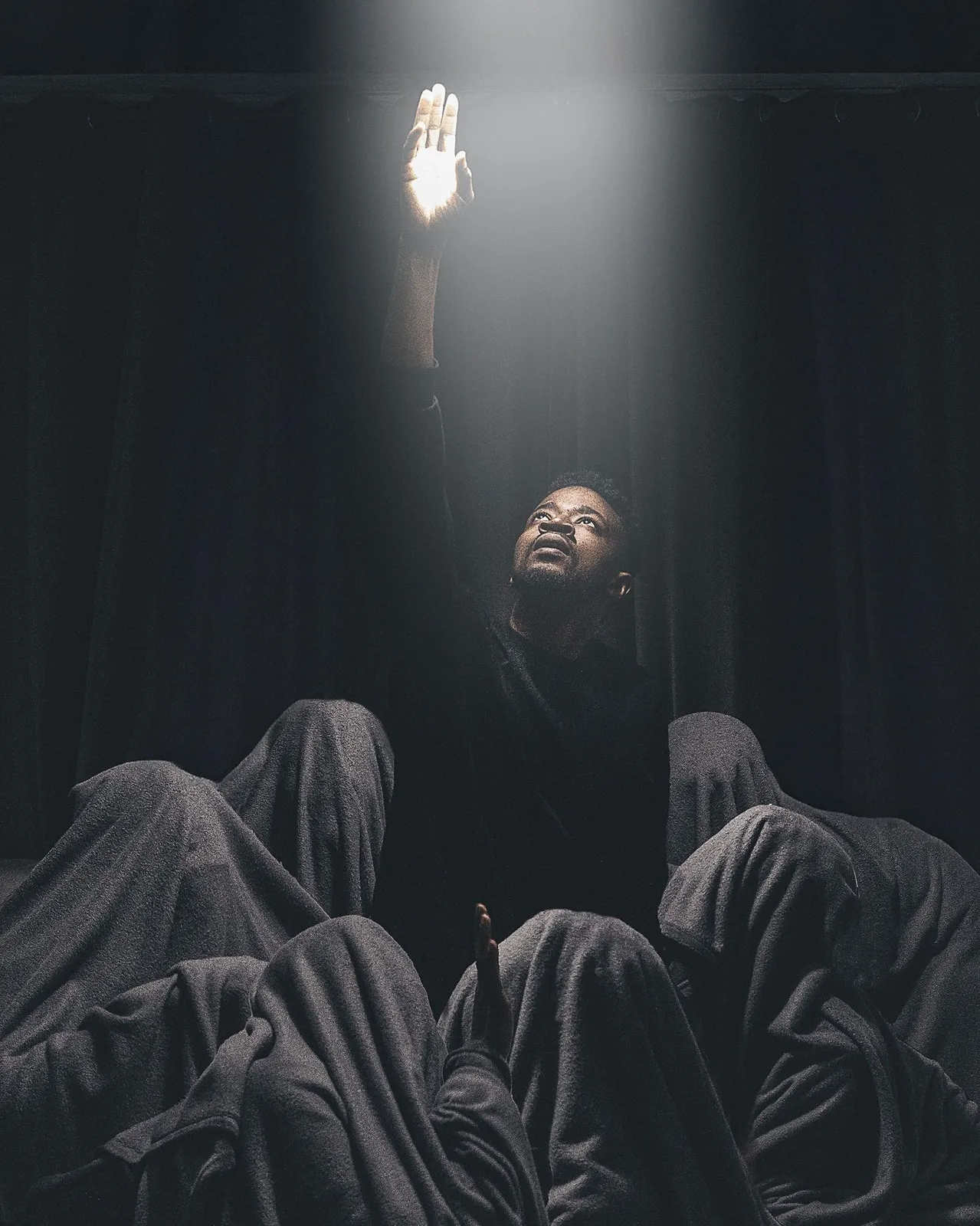 A dramatic artistic composition with dark figures in black robes beneath a single bright light source above.