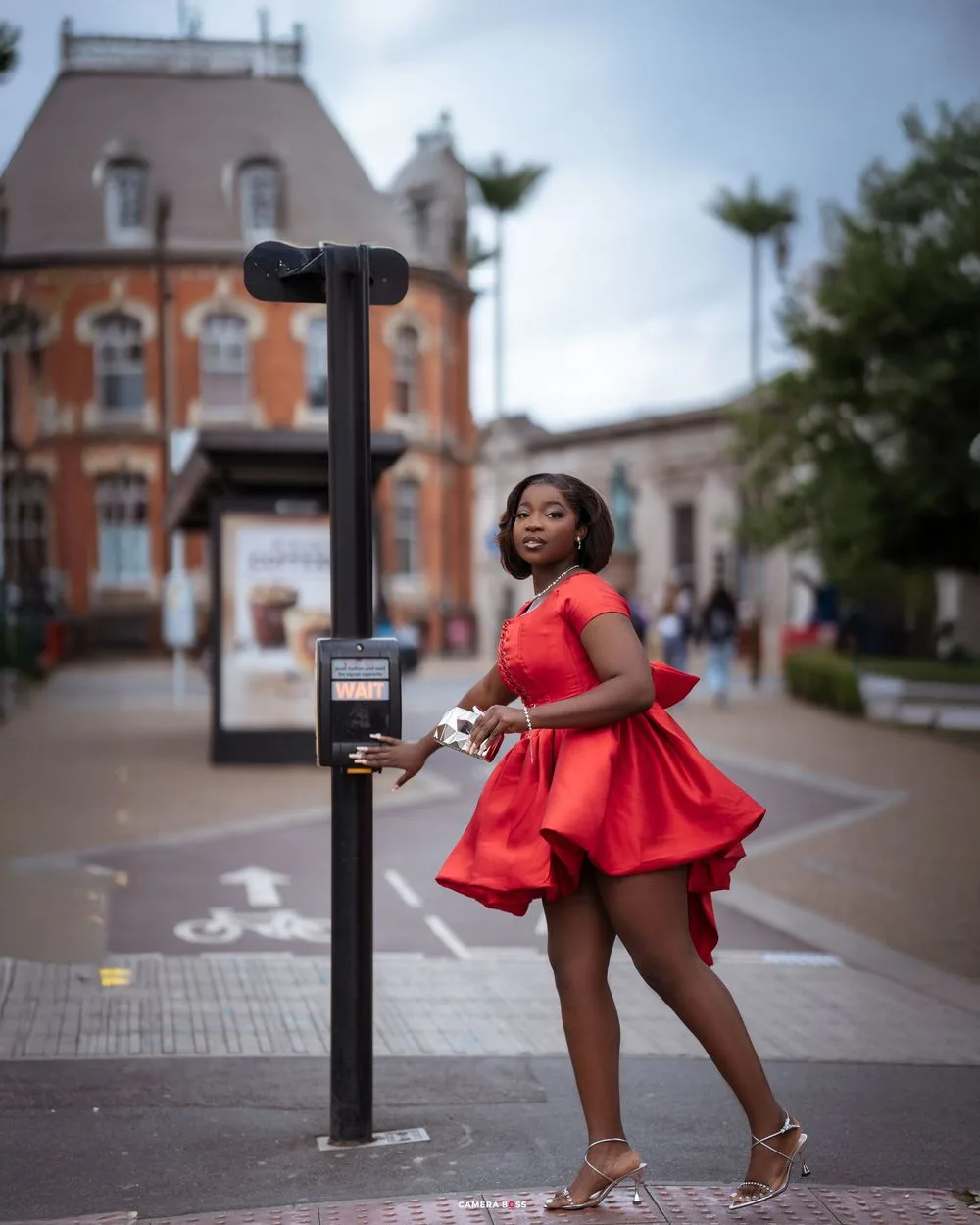 Person in a red dress poses playfully near a parking meter on a city sidewalk with brick buildings behind.