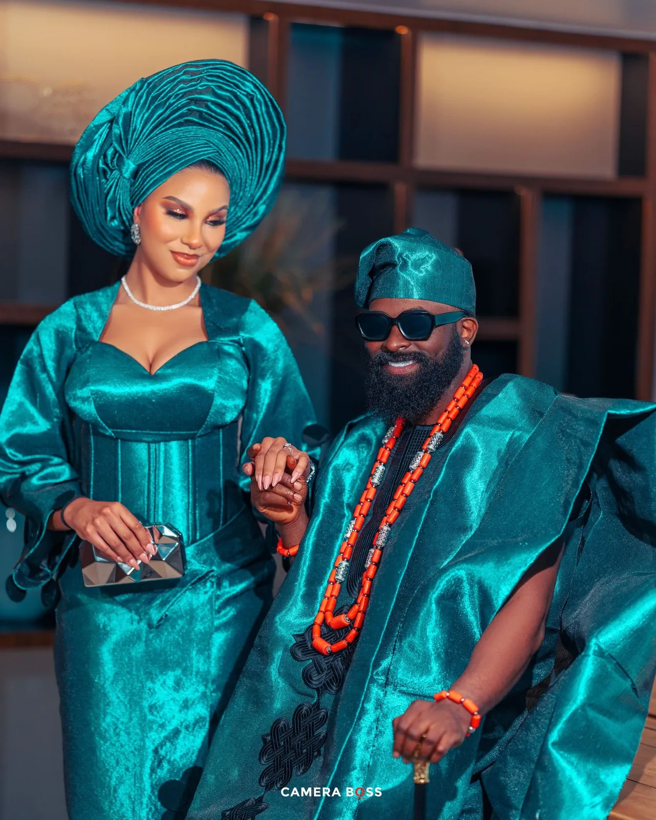 A couple wearing matching teal traditional African attire with a coral beaded necklace pose together at an event.