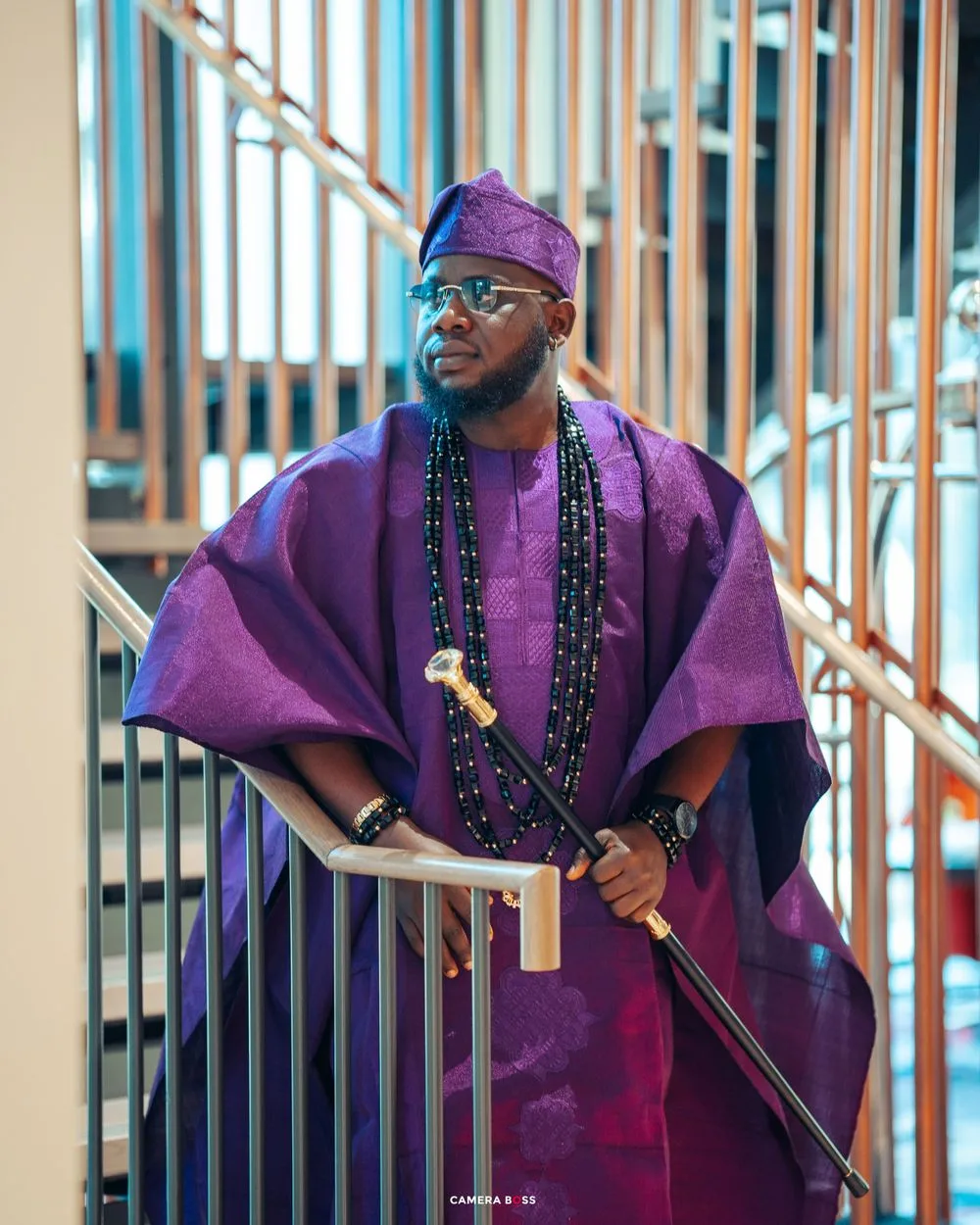 Traditional African attire in rich purple agbada with beaded necklaces and a ceremonial staff against metal railings.