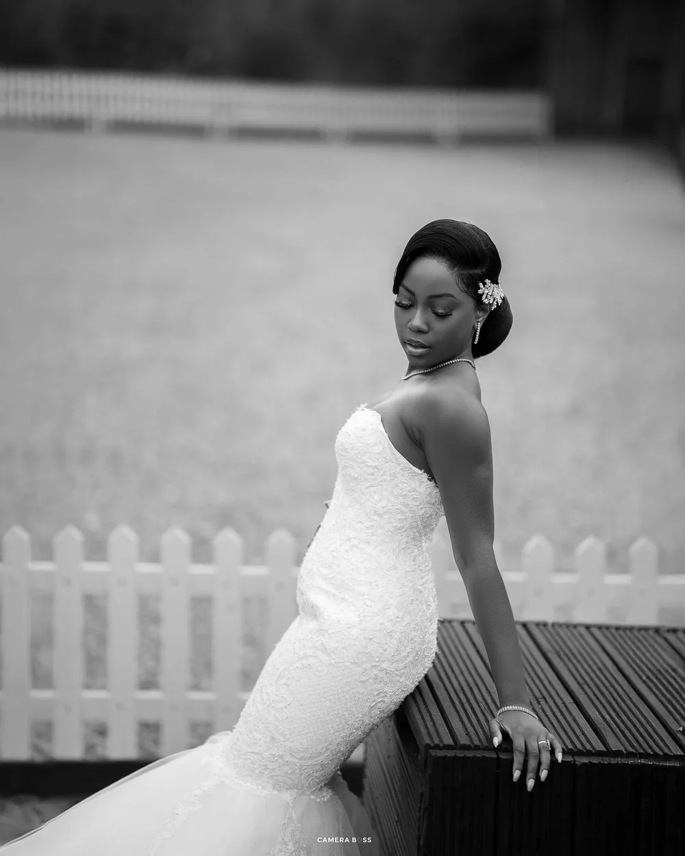 Elegant black and white bridal portrait showing a bride in a fitted lace dress posing beside a white picket fence.
