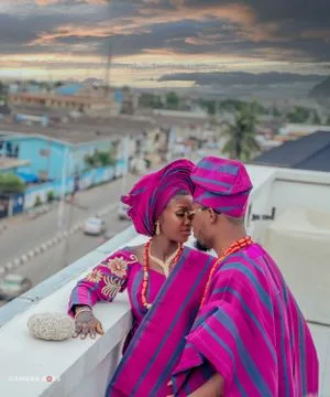 A couple in vibrant pink traditional African attire share an intimate moment on a rooftop at sunset.