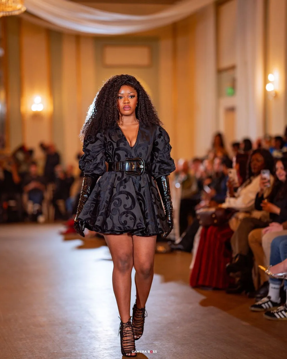 Model walks runway in black textured mini dress with voluminous sleeves and belt during elegant fashion show.