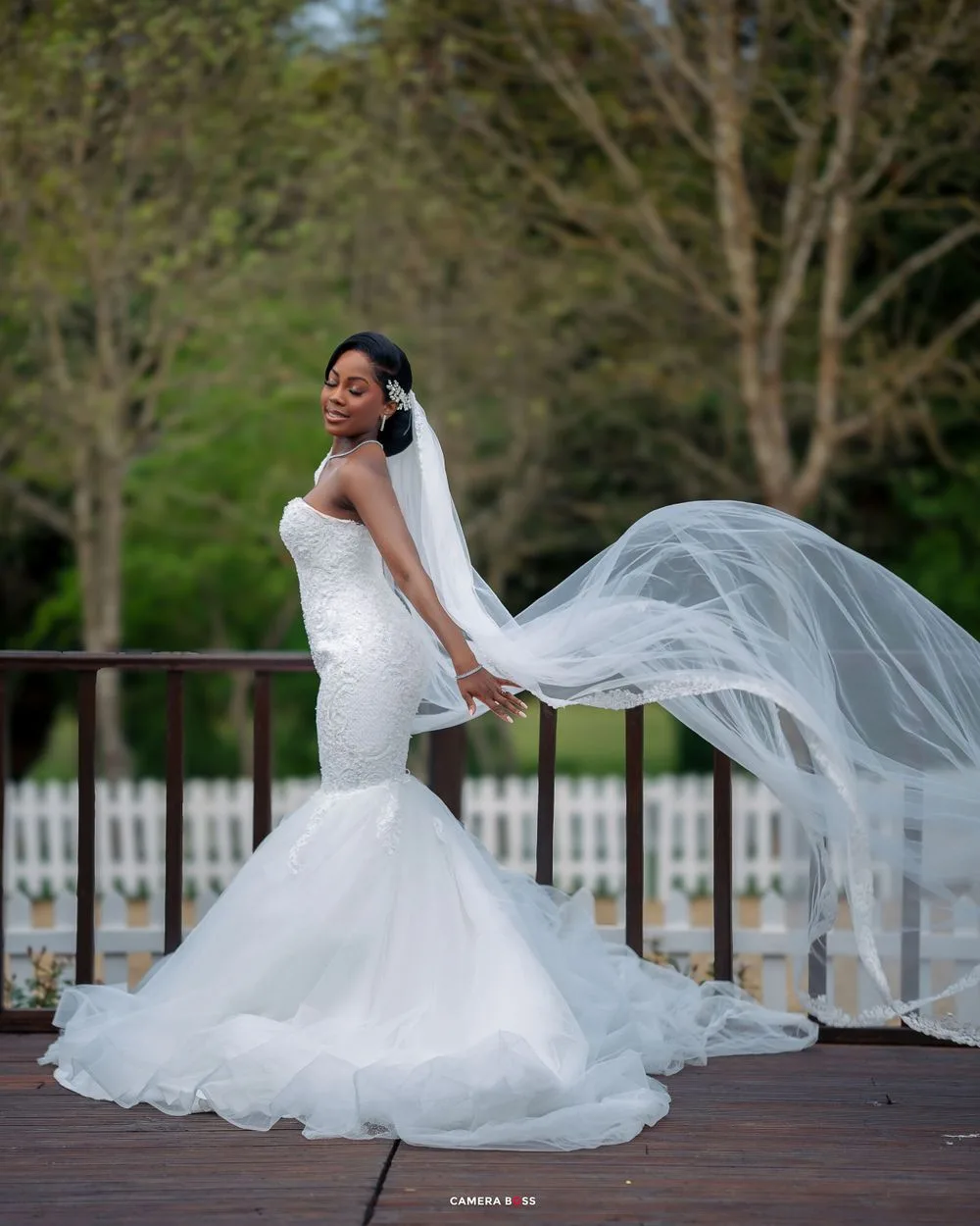 A stunning bridal portrait with a flowing cathedral veil billowing in the wind on a wooden deck surrounded by greenery.