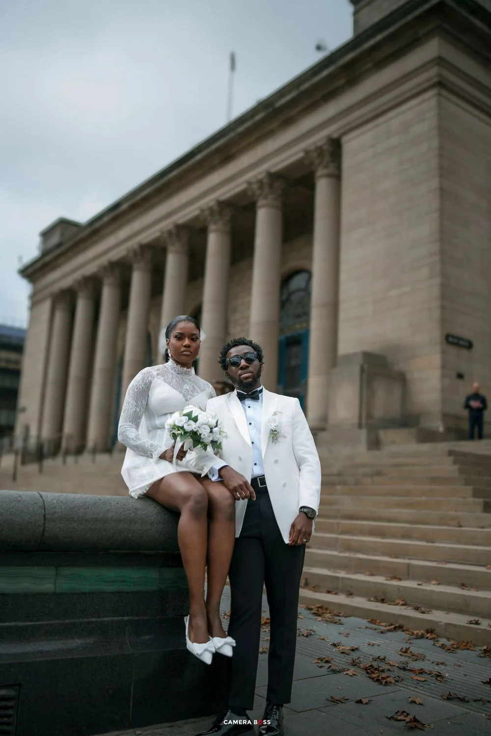 A bride in a white mini dress and groom in a white tuxedo pose at a grand neoclassical building for their wedding photos.