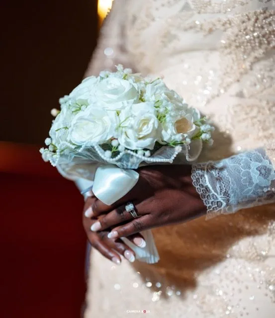 A white rose and baby's breath wedding bouquet held by hands wearing lace sleeves and a diamond ring.