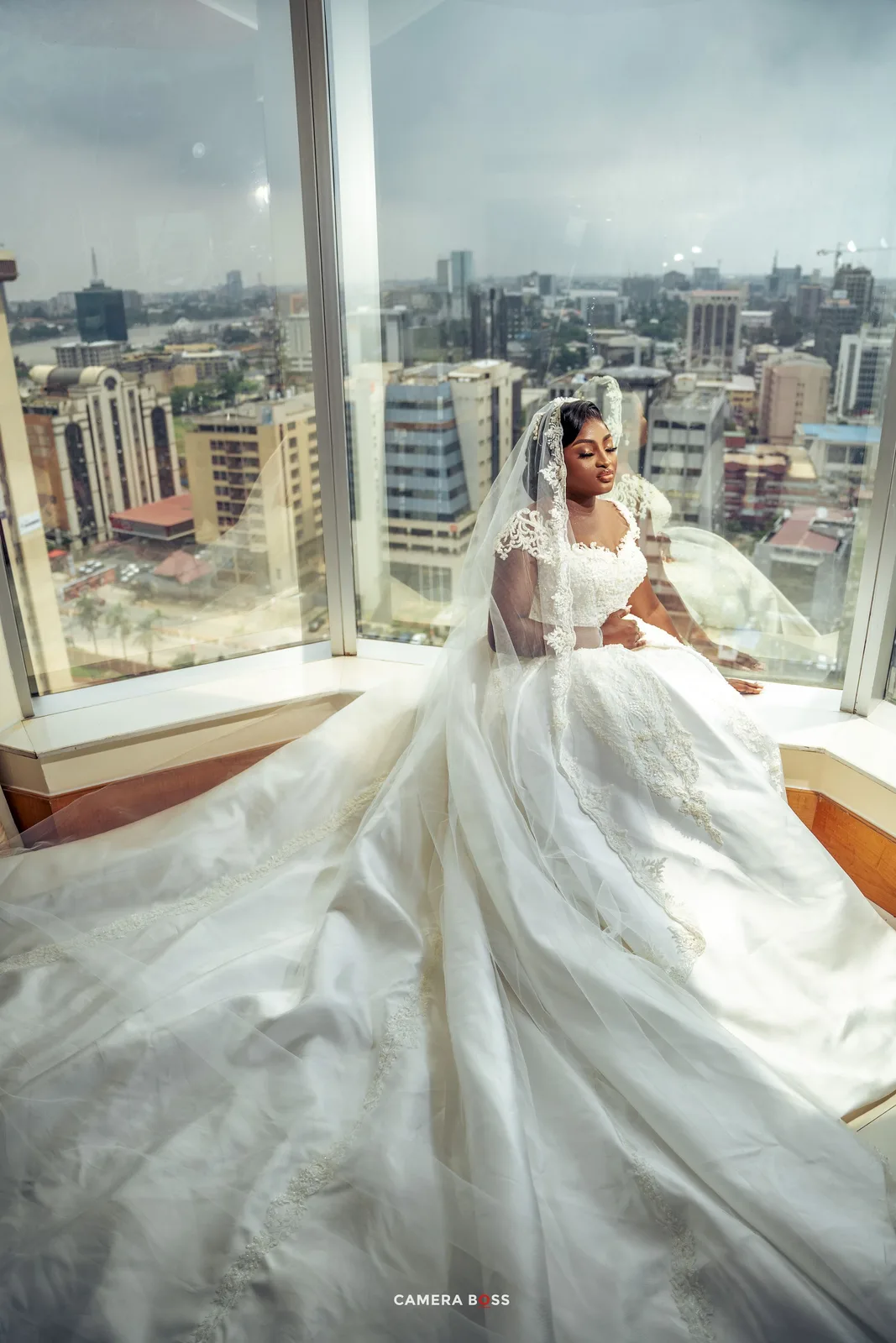 A bride in a flowing white gown poses by floor-to-ceiling windows overlooking a city skyline.